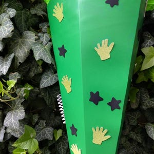Peut inclure: Un c&ocirc;ne de papier vert d&eacute;cor&eacute; de d&eacute;coupes de mains dor&eacute;es paillet&eacute;es et d'&eacute;toiles violet fonc&eacute;. Le c&ocirc;ne est rempli de papier de soie vert. L'arri&egrave;re-plan est un mur de lierre vert.