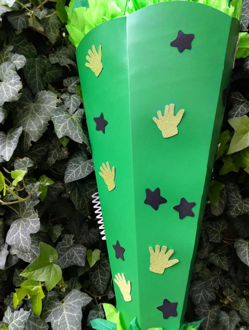 Peut inclure: Un c&ocirc;ne de papier vert d&eacute;cor&eacute; de d&eacute;coupes de mains dor&eacute;es paillet&eacute;es et d'&eacute;toiles violet fonc&eacute;. Le c&ocirc;ne est rempli de papier de soie vert. L'arri&egrave;re-plan est un mur de lierre vert.