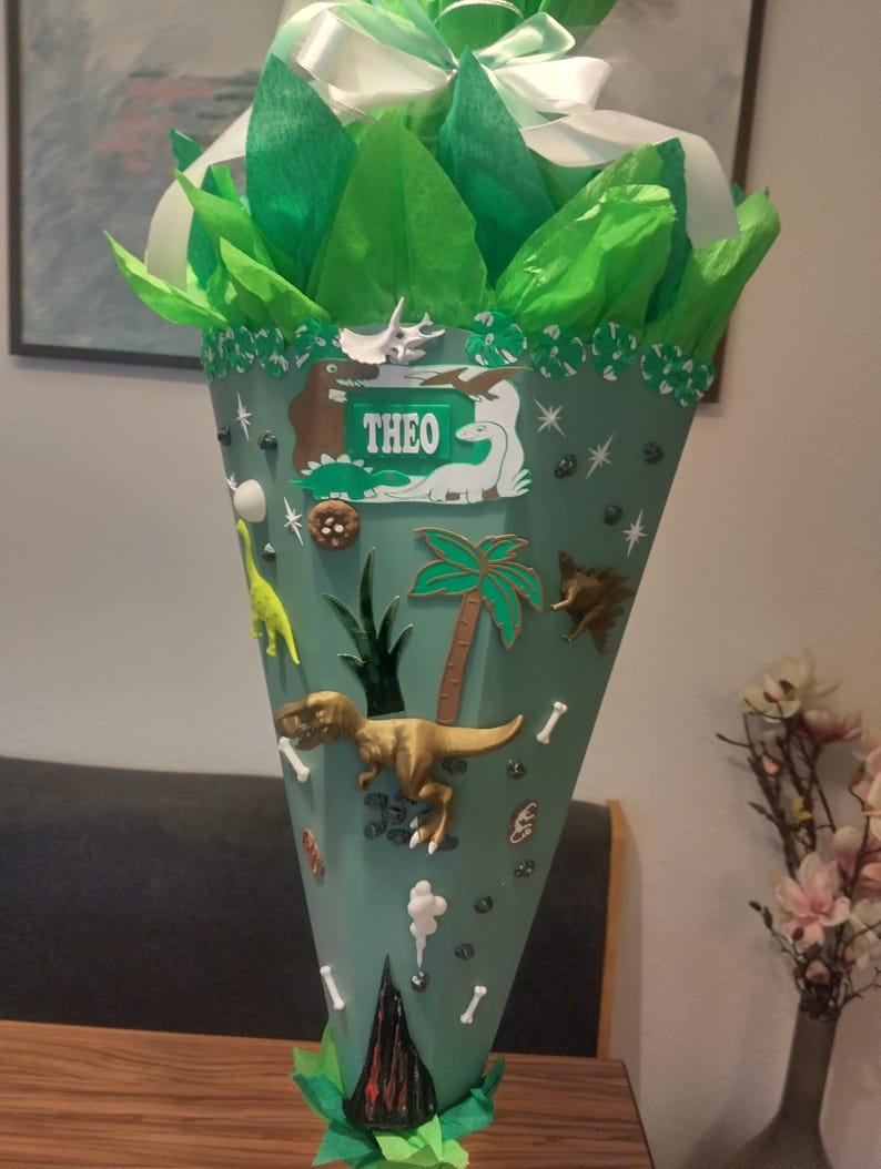 Op de afbeelding: Een kegelvormige schoolcadeauhouder in saliegroen, versierd met diverse dinosaurusfiguren, palmbomen en botvormen. De naam THEO staat op een rechthoekig label bij de bovenkant. De bovenkant is gevuld met felgroen vloeipapier.