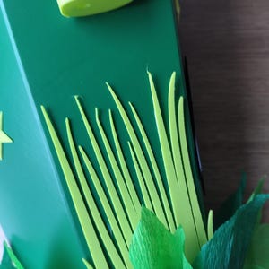Peut inclure: Gros plan sur une bo&icirc;te verte d&eacute;cor&eacute;e d'herbe vert clair et de feuilles vert fonc&eacute;. Un objet cylindrique vert clair est visible en haut de la bo&icirc;te. La bo&icirc;te est sur une surface en bois.