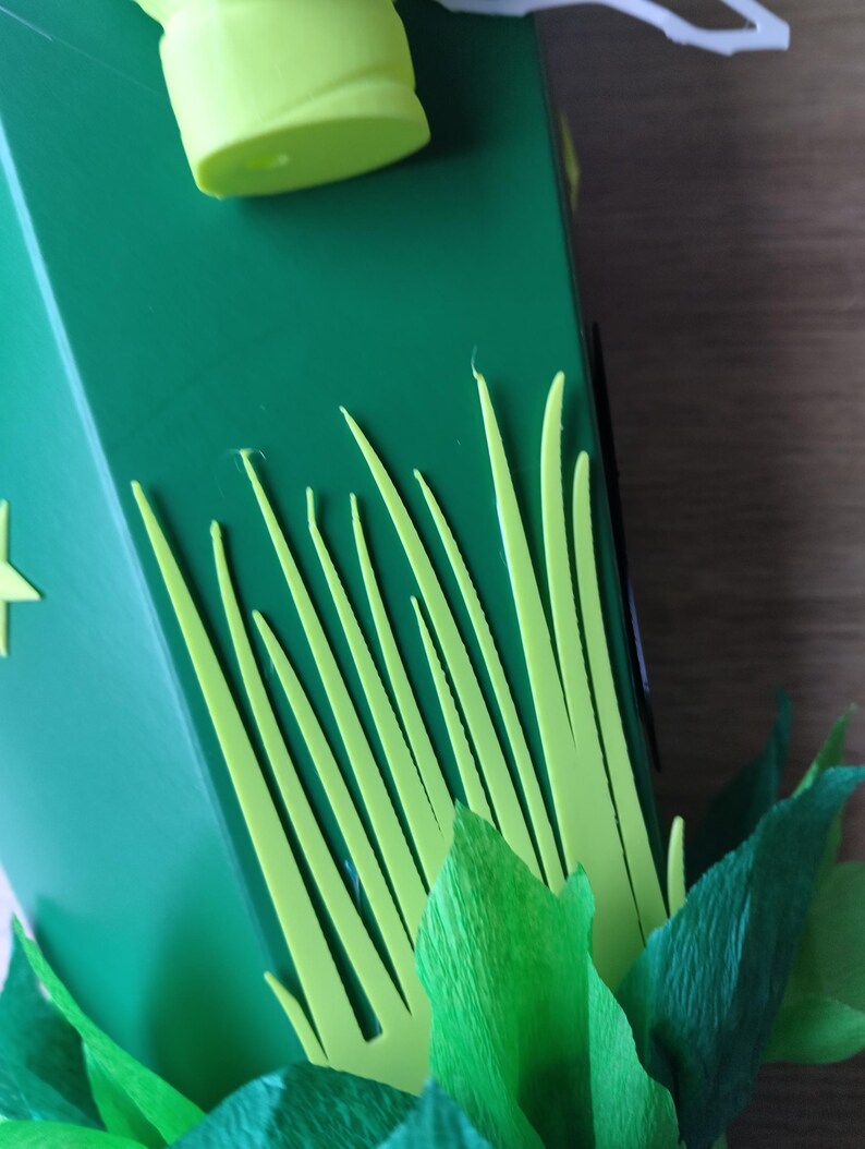Peut inclure: Gros plan sur une bo&icirc;te verte d&eacute;cor&eacute;e d'herbe vert clair et de feuilles vert fonc&eacute;. Un objet cylindrique vert clair est visible en haut de la bo&icirc;te. La bo&icirc;te est sur une surface en bois.