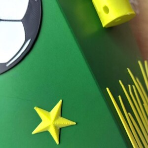 Peut inclure: Une bo&icirc;te verte avec un motif de ballon de football et une &eacute;toile jaune. La bo&icirc;te a un ballon de football sur le c&ocirc;t&eacute; et des accents jaunes. L'&eacute;toile est de couleur jaune vif.
