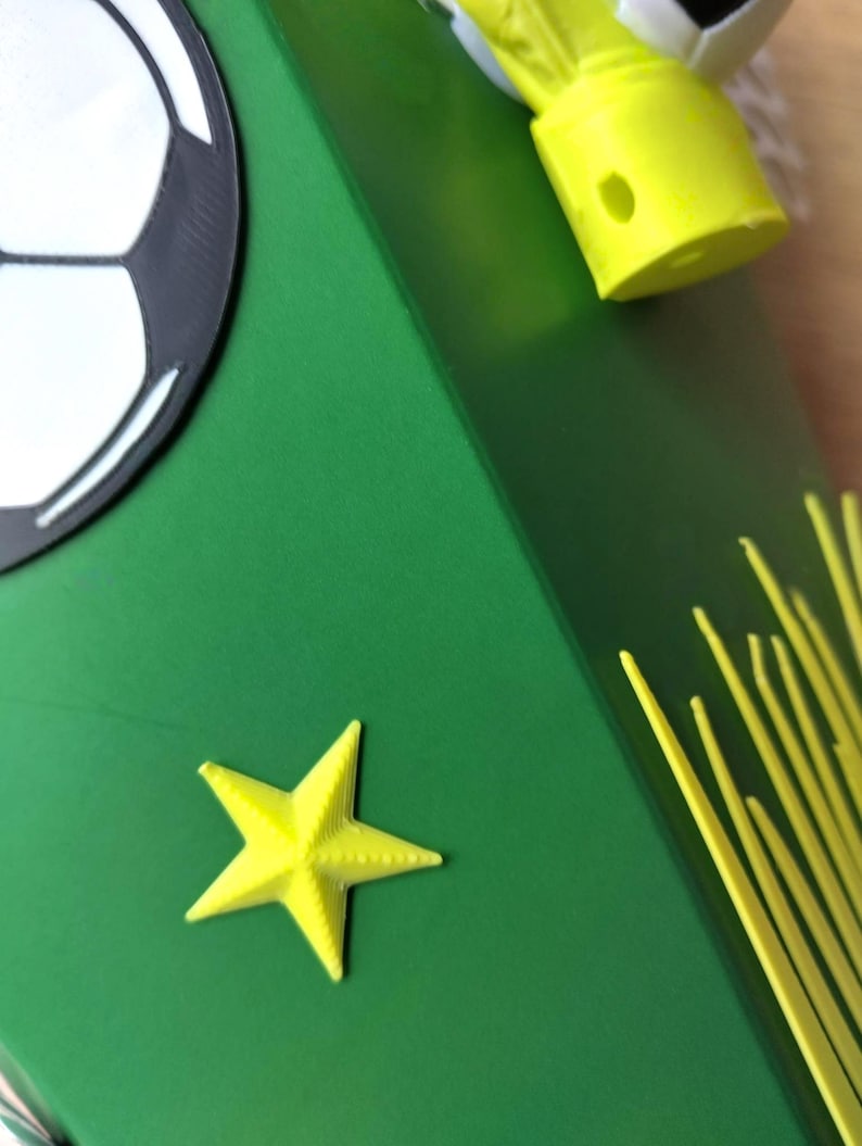 Peut inclure: Une bo&icirc;te verte avec un motif de ballon de football et une &eacute;toile jaune. La bo&icirc;te a un ballon de football sur le c&ocirc;t&eacute; et des accents jaunes. L'&eacute;toile est de couleur jaune vif.
