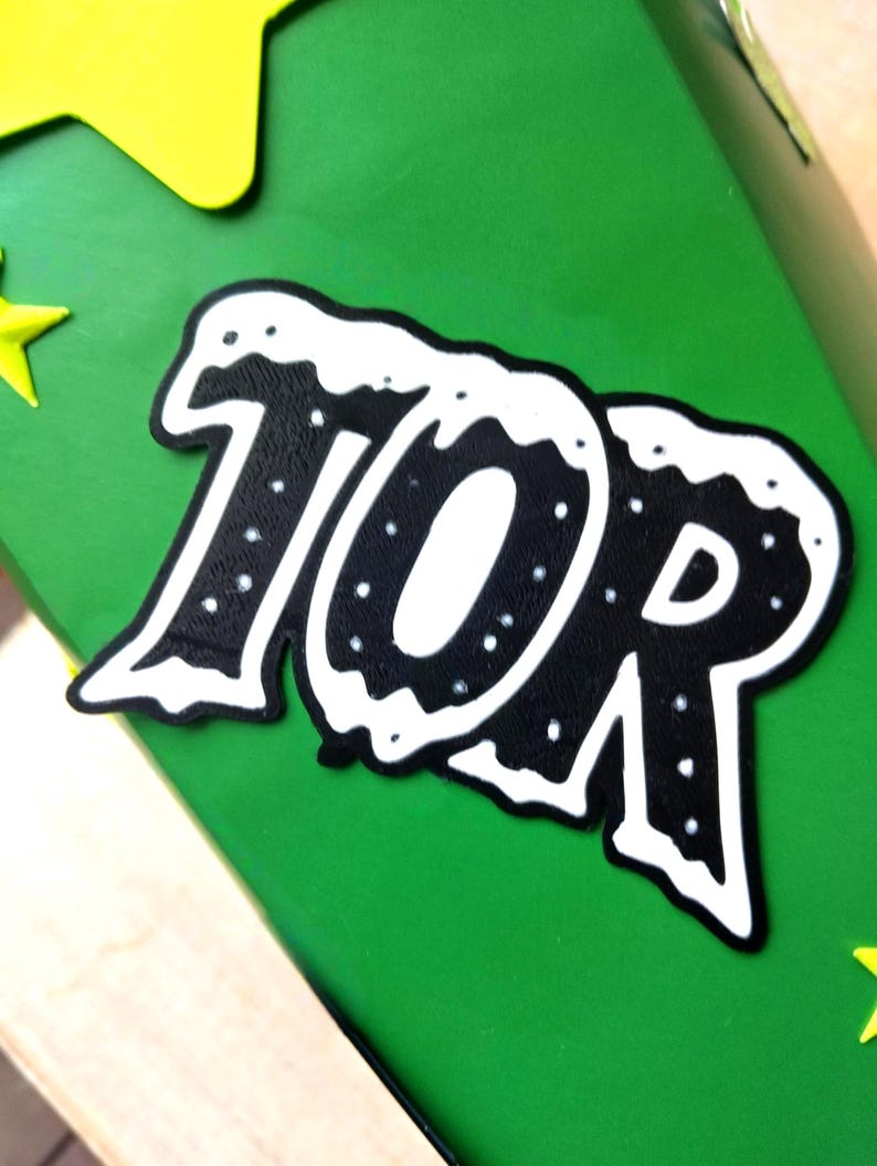 Peut inclure: Gros plan sur une surface verte avec un graphisme noir et blanc indiquant "10R". Le graphisme a une bordure blanche et des points blancs. Des accents en forme d'&eacute;toiles jaunes sont visibles dans les coins.
