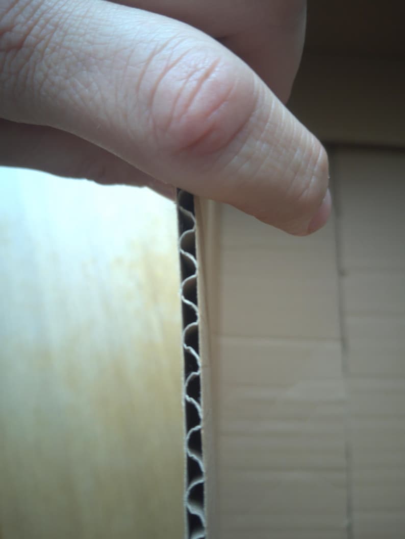 Peut inclure: Gros plan sur du carton ondul&eacute; montrant la construction en couches. Le bord r&eacute;v&egrave;le la structure int&eacute;rieure ondul&eacute;e, avec une main tenant le carton. Le carton est de couleur brun clair.