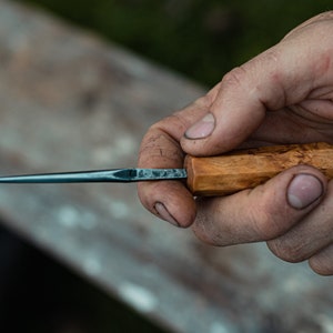 May include: A hand holds a small, sharp knife with a wooden handle. The blade is dark blue and the handle is light brown with a natural wood grain.