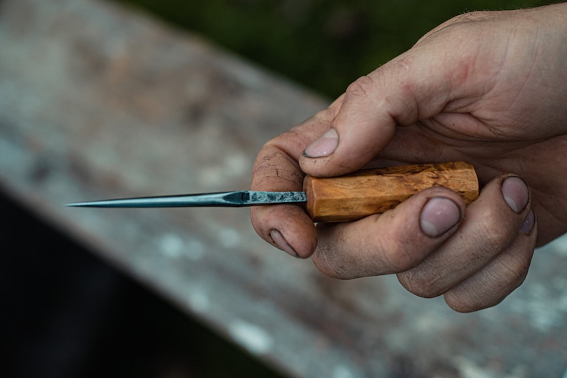 May include: A hand holds a small, sharp knife with a wooden handle. The blade is dark blue and the handle is light brown with a natural wood grain.