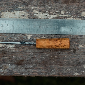 May include: A small, wooden-handled tool with a thin, pointed metal blade. The tool is lying on a wooden surface next to a metal ruler marked in millimeters.