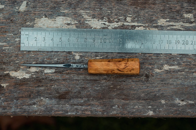 May include: A small, wooden-handled tool with a thin, pointed metal blade. The tool is lying on a wooden surface next to a metal ruler marked in millimeters.