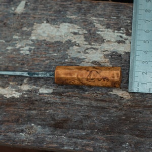 May include: A small, wooden-handled knife with a metal blade. The handle is brown and has a wood grain pattern. The blade is about 5 inches long and is sharp. The knife is lying on a wooden surface next to a ruler.