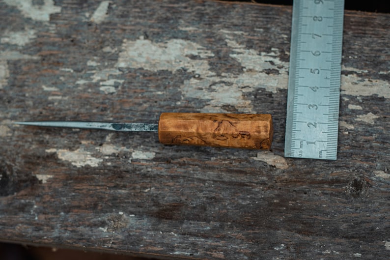 May include: A small, wooden-handled knife with a metal blade. The handle is brown and has a wood grain pattern. The blade is about 5 inches long and is sharp. The knife is lying on a wooden surface next to a ruler.