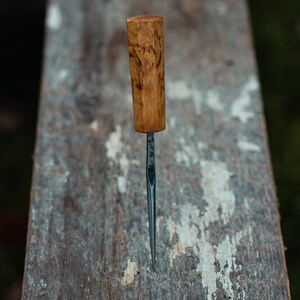 May include: A close-up of a wooden-handled carving tool with a thin, sharp metal blade. The tool is resting on a weathered wooden surface.