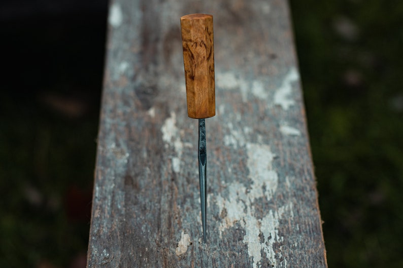 May include: A close-up of a wooden-handled carving tool with a thin, sharp metal blade. The tool is resting on a weathered wooden surface.