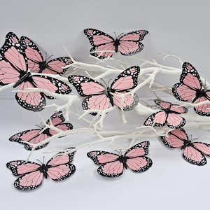 Puede incluir: Una colección de decoraciones de mariposas rosas y negras con detalles blancos. Las mariposas están dispuestas sobre ramas blancas, creando una exhibición caprichosa y decorativa. Las mariposas tienen patrones de alas detallados.