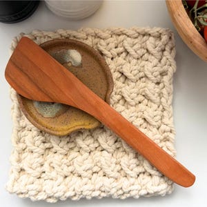 May include: A wooden spatula rests on a ceramic spoon rest, atop a cream-colored, woven trivet. The spatula and spoon rest are brown, and the trivet has a textured, braided pattern. A wooden bowl with red tomatoes is in the background.