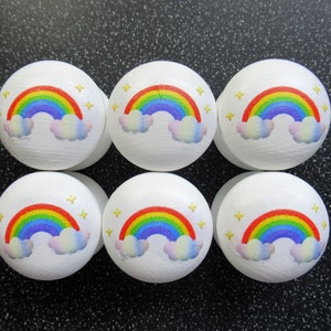 May include: Six white wooden knobs with rainbow and cloud designs. Each knob features a rainbow with red, orange, yellow, green, blue, indigo, and violet stripes. The clouds are white with a light pink and blue gradient. Each knob has small yellow stars scattered around the design.