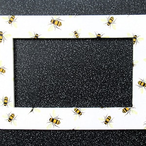 May include: A white picture frame with a border of yellow and black bees. The frame is made of wood and has a white background.