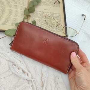Puede incluir: Una cartera de cuero marrón con cremallera y un logotipo "LB" en relieve dorado. La cartera está sobre una superficie de crochet blanco.