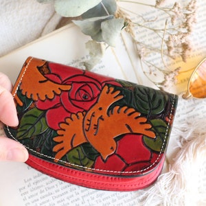 Puede incluir: Una cartera de cuero roja con un diseño floral con rosas y pájaros. La cartera tiene una forma curva y una puntada blanca alrededor del borde.