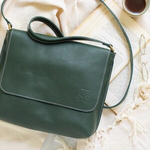 May include: A dark green leather crossbody bag with a white stitch detail and a small embossed logo. The bag is on a white fabric background.