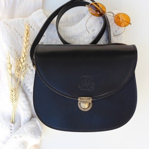 May include: A black leather satchel bag with a brass clasp and an engraved letter "L". The bag has a long strap and is on a white surface.