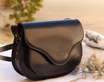 May include: A black leather satchel bag with a flap closure and a gold metal buckle. The bag has a monogrammed "HB" on the front.