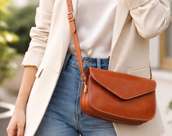 May include: A woman wearing a cream-colored blazer, white top, and blue jeans, with a brown leather crossbody bag. The bag has a flap closure and a gold-colored buckle. The bag's strap is also brown leather.