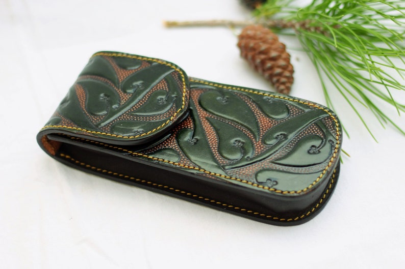May include: A green leather case with a floral design and yellow stitching. The case is closed and has a flap.
