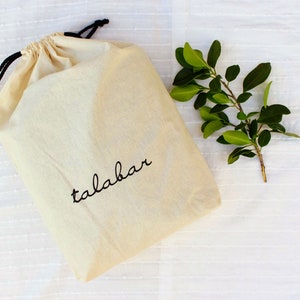 May include: A white drawstring bag with black drawstrings and the word "talabar" embroidered in black on the front. The bag is lying on a white textured surface with a small green plant to the right.