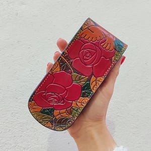 Op de afbeelding: Een rode leren portemonnee met een bloemenontwerp met rozen en bladeren. De portemonnee is gegraveerd met een gedetailleerd patroon en heeft gele stiksels rond de randen.