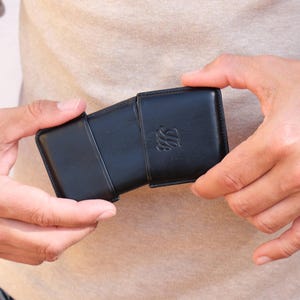 May include: A black leather case with a flap closure, held open to reveal the interior. The case has a debossed emblem on the front. The case is rectangular and appears to be designed to hold a small item.