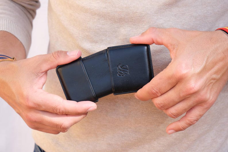 May include: A black leather case with a flap closure, held open to reveal the interior. The case has a debossed emblem on the front. The case is rectangular and appears to be designed to hold a small item.