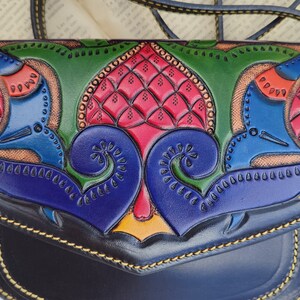May include: A handcrafted leather crossbody bag with a colorful, embossed design. The bag features a flap closure and a long strap. The design includes red, blue, green, and yellow accents, with intricate patterns and stitching.