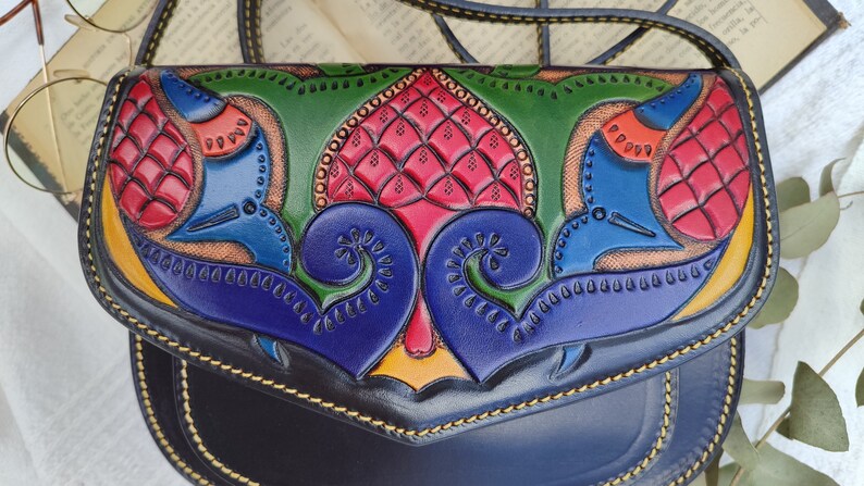 May include: A handcrafted leather crossbody bag with a colorful, embossed design. The bag features a flap closure and a long strap. The design includes red, blue, green, and yellow accents, with intricate patterns and stitching.