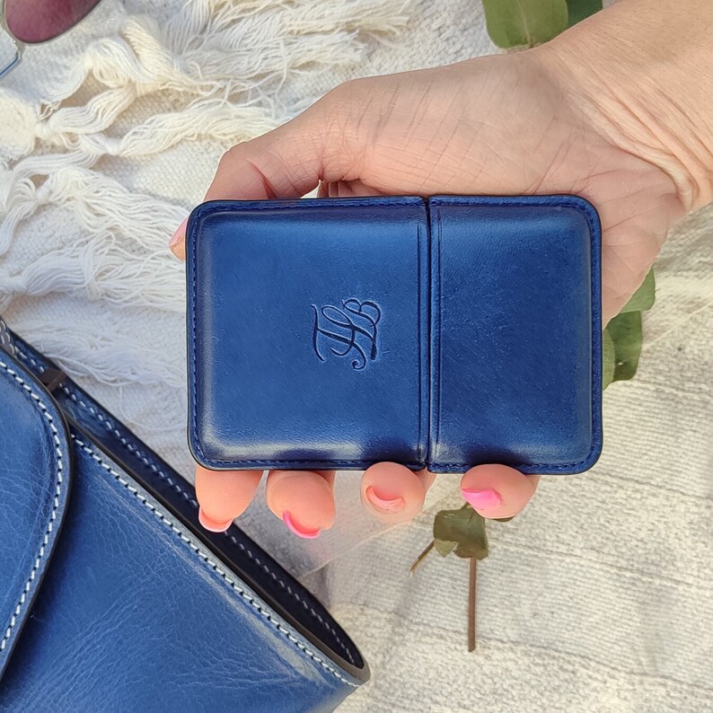 May include: A navy blue leather case with a debossed monogram. The case is rectangular with rounded corners and a hinged design. The case is held in a hand, with a matching blue purse visible in the background.