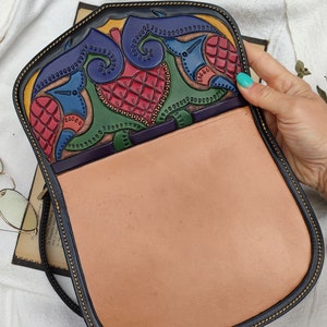 May include: A handcrafted leather shoulder bag with a colorful, intricate design on the flap. The interior is a light tan color, and the bag has a black border with yellow stitching. The flap features red, blue, green, and yellow patterns.