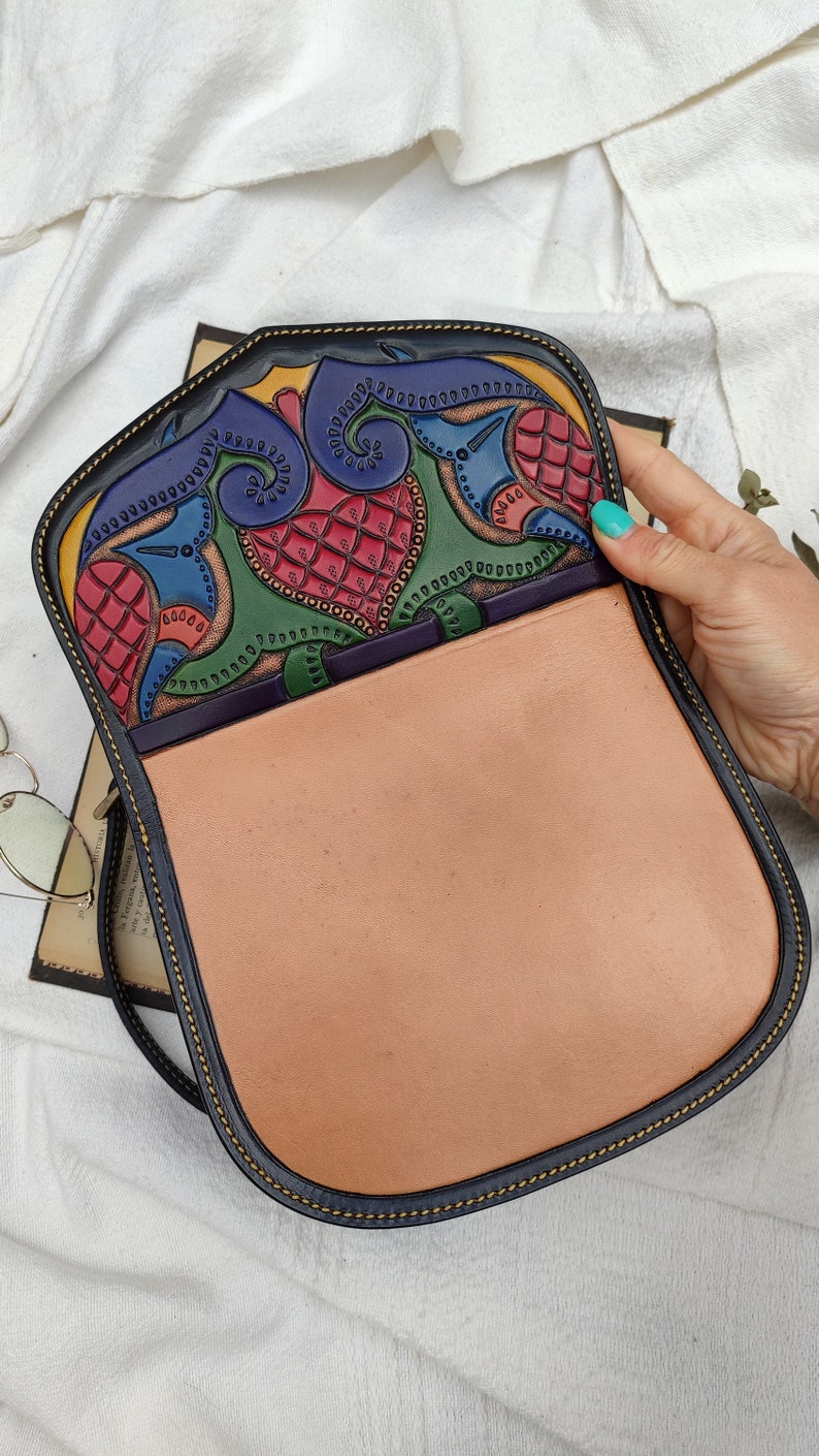 May include: A handcrafted leather shoulder bag with a colorful, intricate design on the flap. The interior is a light tan color, and the bag has a black border with yellow stitching. The flap features red, blue, green, and yellow patterns.