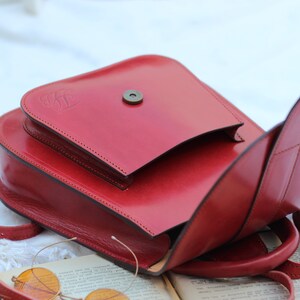 May include: A red leather satchel bag with a flap closure and a small metal button. The bag has a strap and is open to reveal the inside.