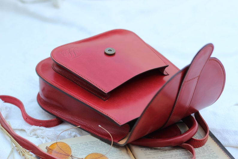 May include: A red leather satchel bag with a flap closure and a small metal button. The bag has a strap and is open to reveal the inside.
