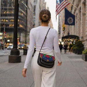 May include: Black leather crossbody bag with a colorful, patterned flap. The bag has a long, adjustable strap and is worn over the shoulder. The bag is carried by a person walking down a city street.