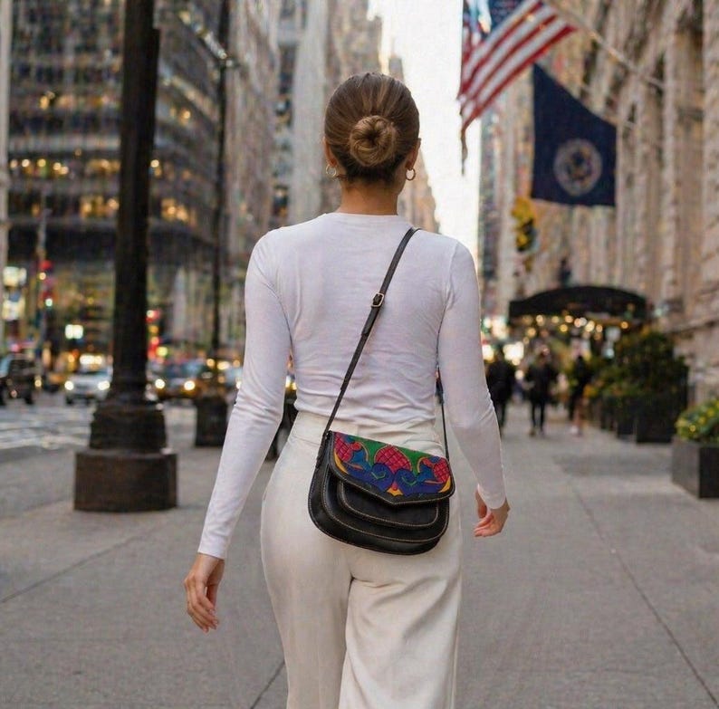 May include: Black leather crossbody bag with a colorful, patterned flap. The bag has a long, adjustable strap and is worn over the shoulder. The bag is carried by a person walking down a city street.