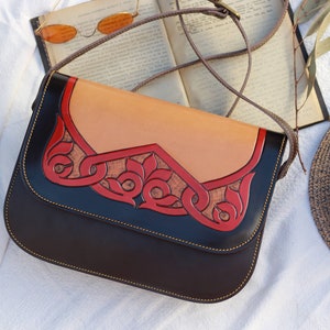 Puede incluir: Un bolso de hombro de cuero marrón con una solapa de cuero tostado y detalles de cuero rojo. El bolso tiene un diseño decorativo en la solapa.