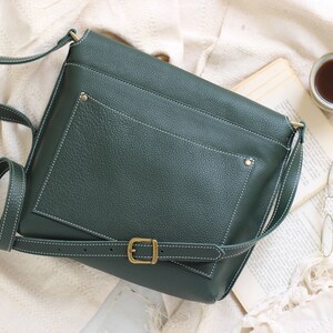 May include: A dark green leather crossbody bag with a white stitch detail and a brass buckle. The bag has a flap closure and a small pocket on the front.