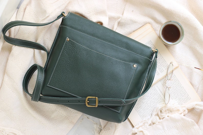 May include: A dark green leather crossbody bag with a white stitch detail and a brass buckle. The bag has a flap closure and a small pocket on the front.