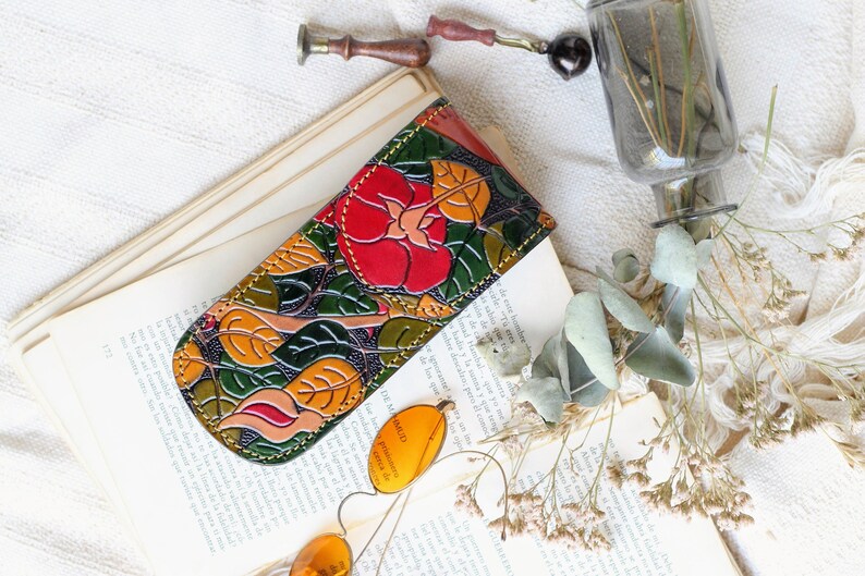 Op de afbeelding: Een leren bril etui met een bloemenpatroon met rode, oranje, gele en groene bladeren. De etui is gesloten en ligt op een boek.