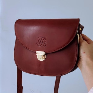 Puede incluir: Un bolso de hombro de cuero burdeos con cierre dorado y un monograma "HB" en la parte delantera.