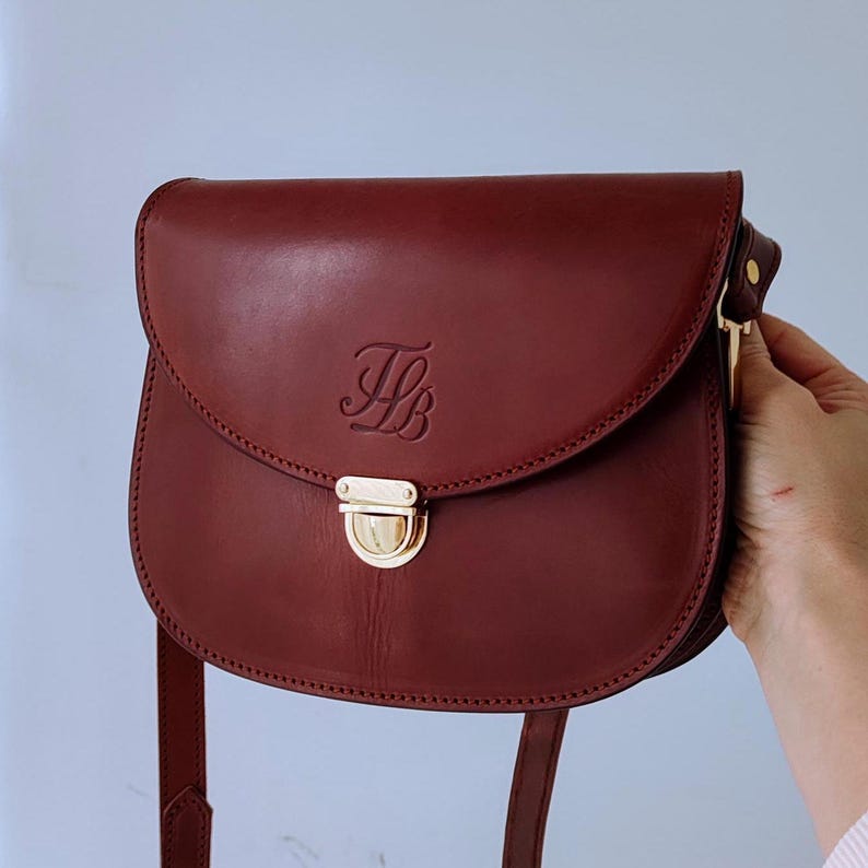 Puede incluir: Un bolso de hombro de cuero burdeos con cierre dorado y un monograma "HB" en la parte delantera.