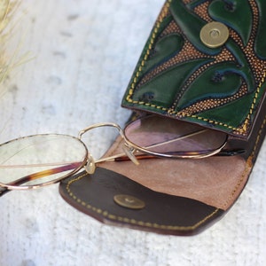May include: A brown leather glasses case with a green floral design and a gold button closure. The case is open and a pair of round, gold-rimmed glasses with clear lenses are inside.