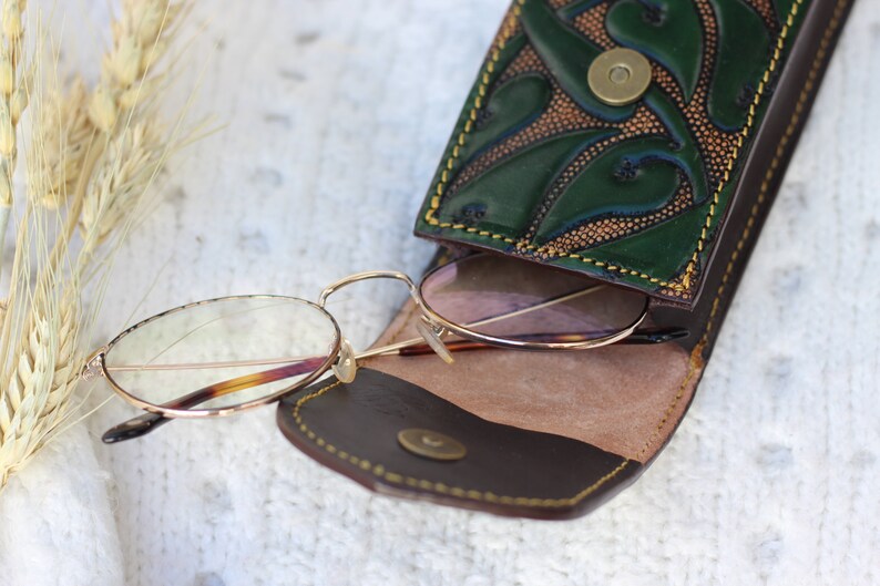 May include: A brown leather glasses case with a green floral design and a gold button closure. The case is open and a pair of round, gold-rimmed glasses with clear lenses are inside.