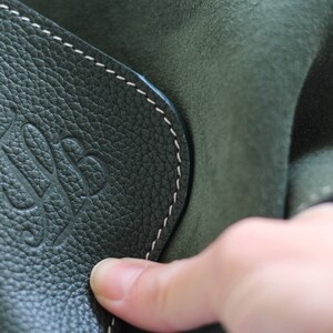May include: Close-up of a dark green leather bag with a white stitched seam and a embossed logo. The logo is a stylized letter 'S' with a leaf design.
